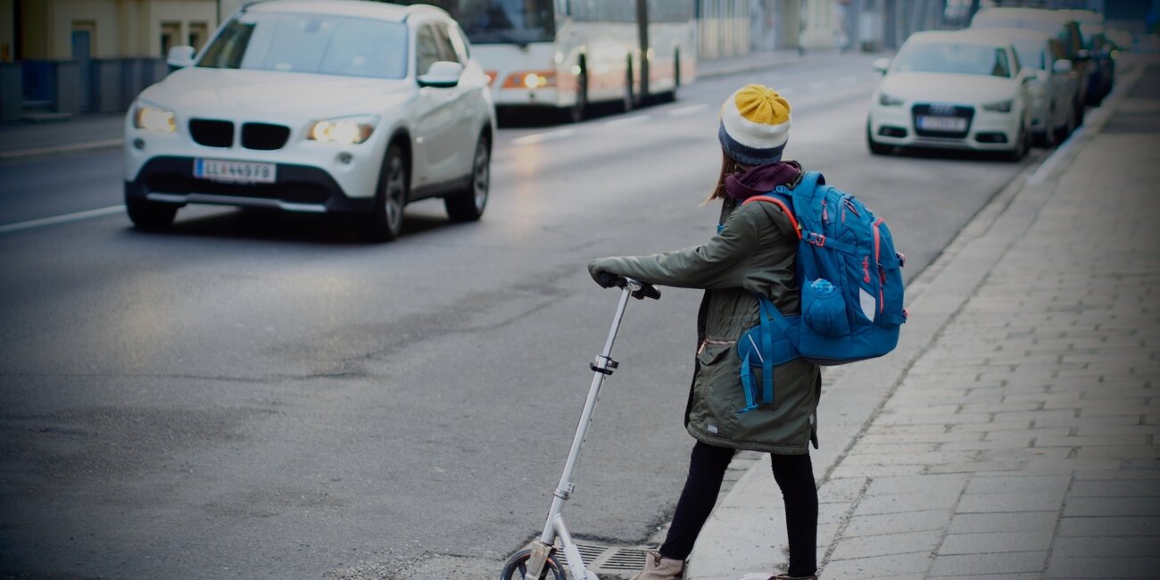Kind mit Rucksack und Tretroller wartet am Fahrbahnrand, auf der Straße sind ein SUV und ein Bus zu sehen