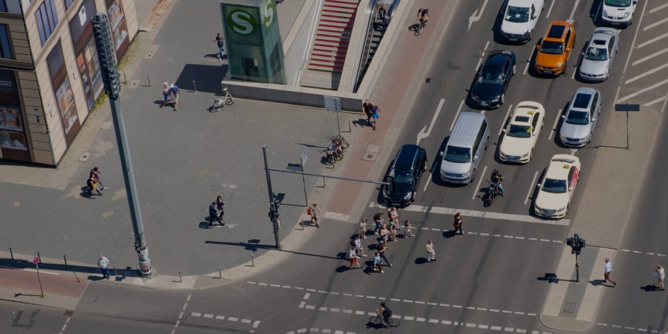 Luftaufnahme einer Kreuzung, an der Autos warten, während Menschen zu Fuß und auf dem Fahrrad die Fahrbahn überqueren; daneben ist der Einganz zu einer S-Bahn-Haltestelle