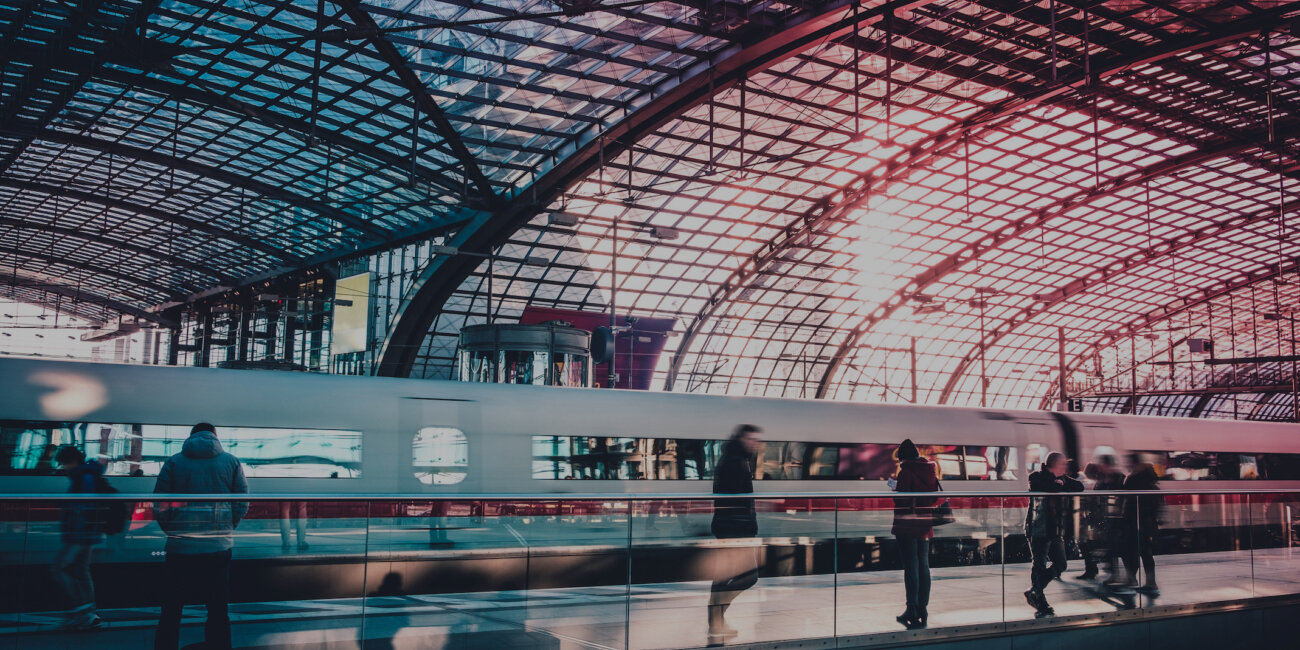 Menschen warten am Berliner Hauptbahnhof auf einen Zug. Im Hintergrund ist ein ICE zu sehen, darüber die wabenartige Dachkonstruktion in blau-rotem Licht.
