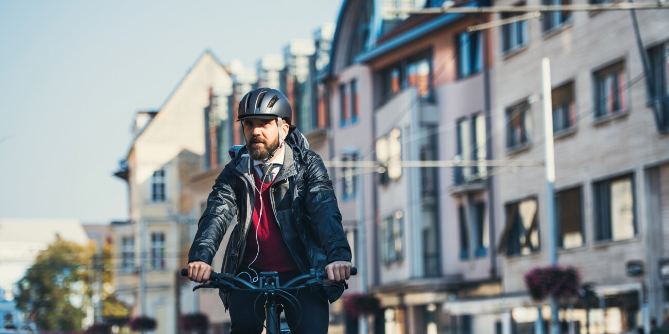 Bärtiger Mann mit Fahrradhelm und Kopfhörern auf einem E-Bike, im Hintergrund ist verschwommen Häuser, Tram-Oberleitungen und parkende Autos zu sehen