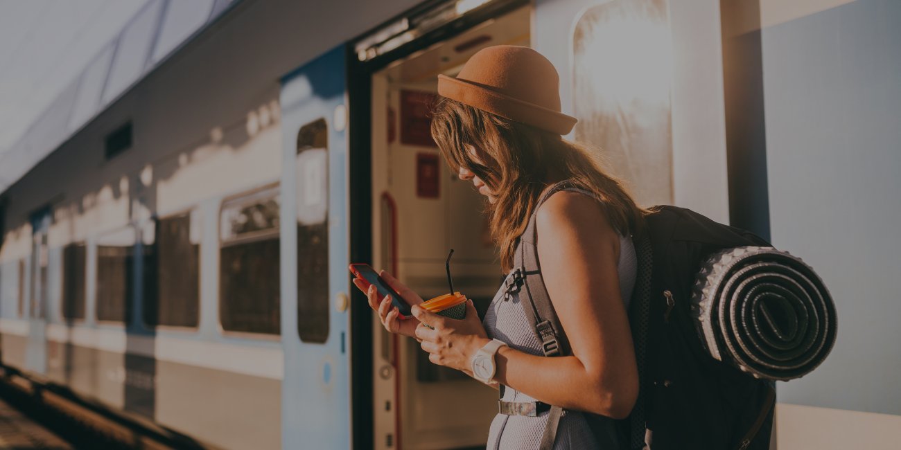Frau mit Reise-Rucksack und Getränk in der Hand steht am Bahnsteig vor einem Zug und schaut auf ihr Smartphone