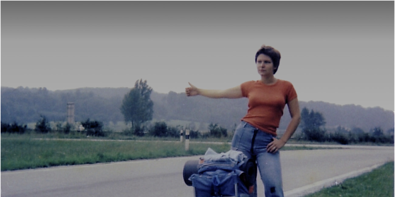 Eine Frau steht mit ausgestrecktem Daumen am Straßenrand, vor ihr ein Rucksack. Sie möchte mitgenommen werden.