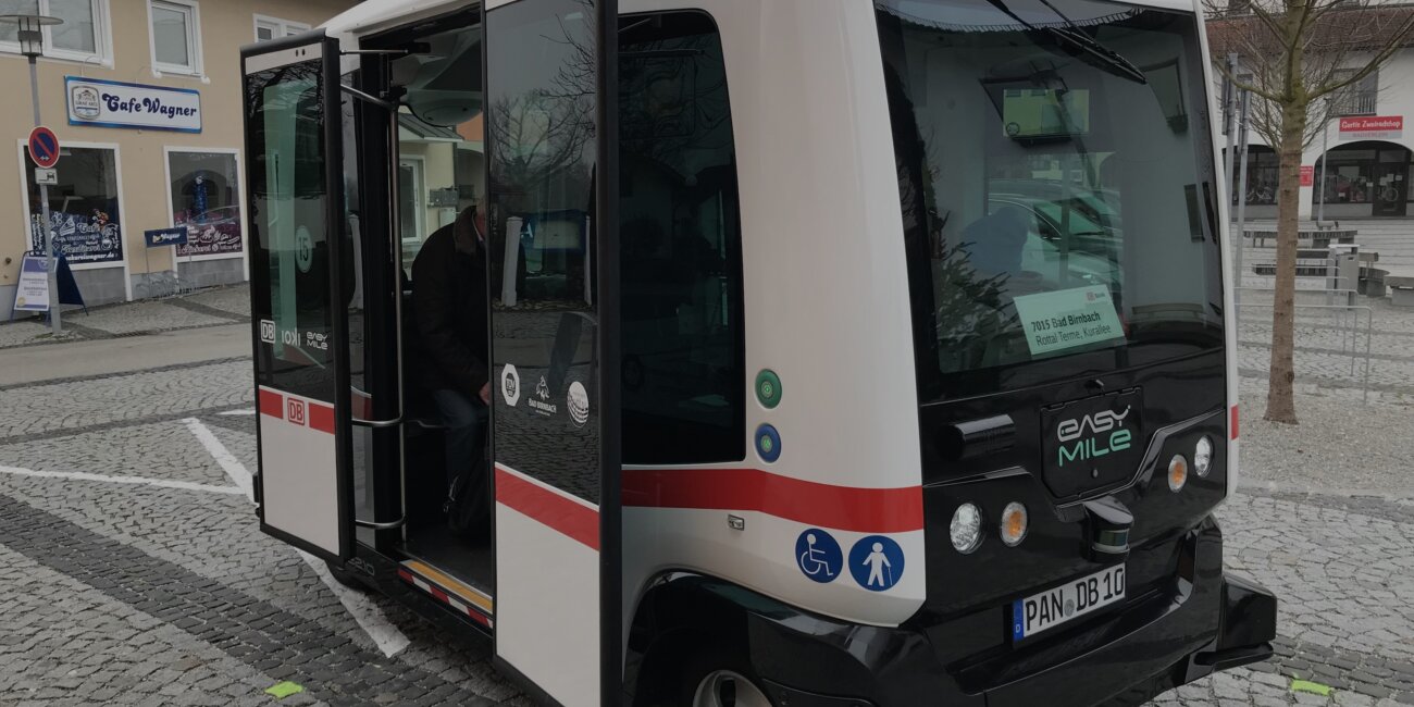 Ein autonom fahrender Kleinbus steht mit geöffneter Tür auf einem Dorfplatz.
