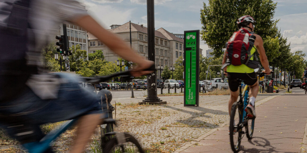 Zwei Menschen fahren auf Rädern. Links neben dem Radweg steht eine Säule mit digitaler Anzeigetafel. Sie zeigt den aktuellen Tageszählerstand vorbeigefahrener Räder an.