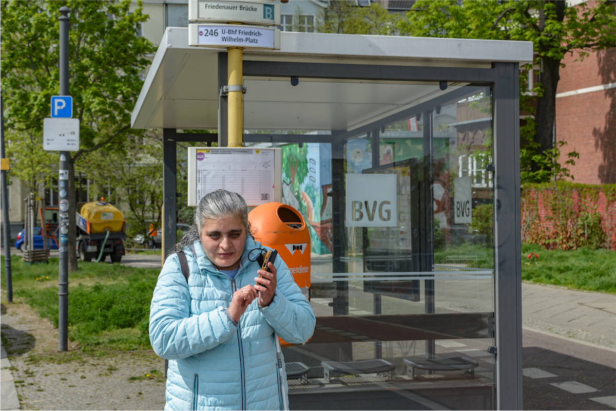 Foto: Eine blinde Frau steht an einer Bushaltestelle und hört ihrem Smartphone zu, dass ihr gerade Audio-Informationen zum Busverkehr übermittelt