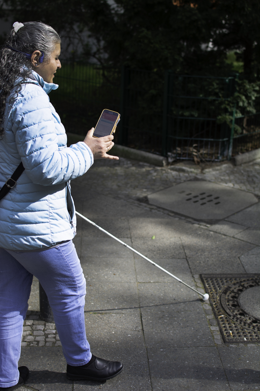 Eine Frau auf dem Gehweg, einen Blindenstock (Langstock) in der einen, ein Smartphone in der anderen Hand