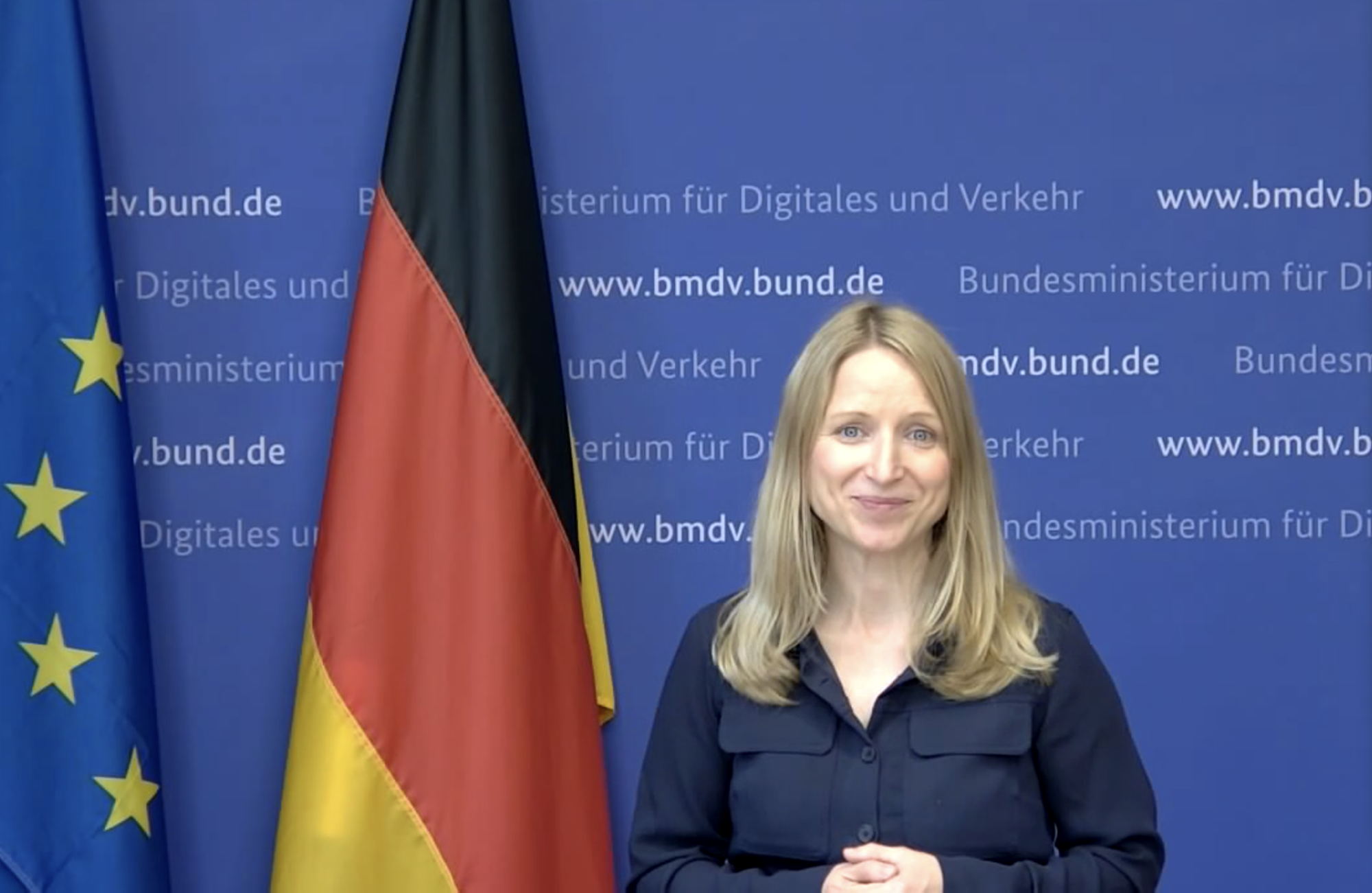 Foto, das Daniela Kluckert zeigt, links neben ihr eine Deutschland-Fahne und eine EU-Fahne, hier ihr eine blaue Wand, auf der die Internetadresse des BMDV mehrfach zu lesen ist.