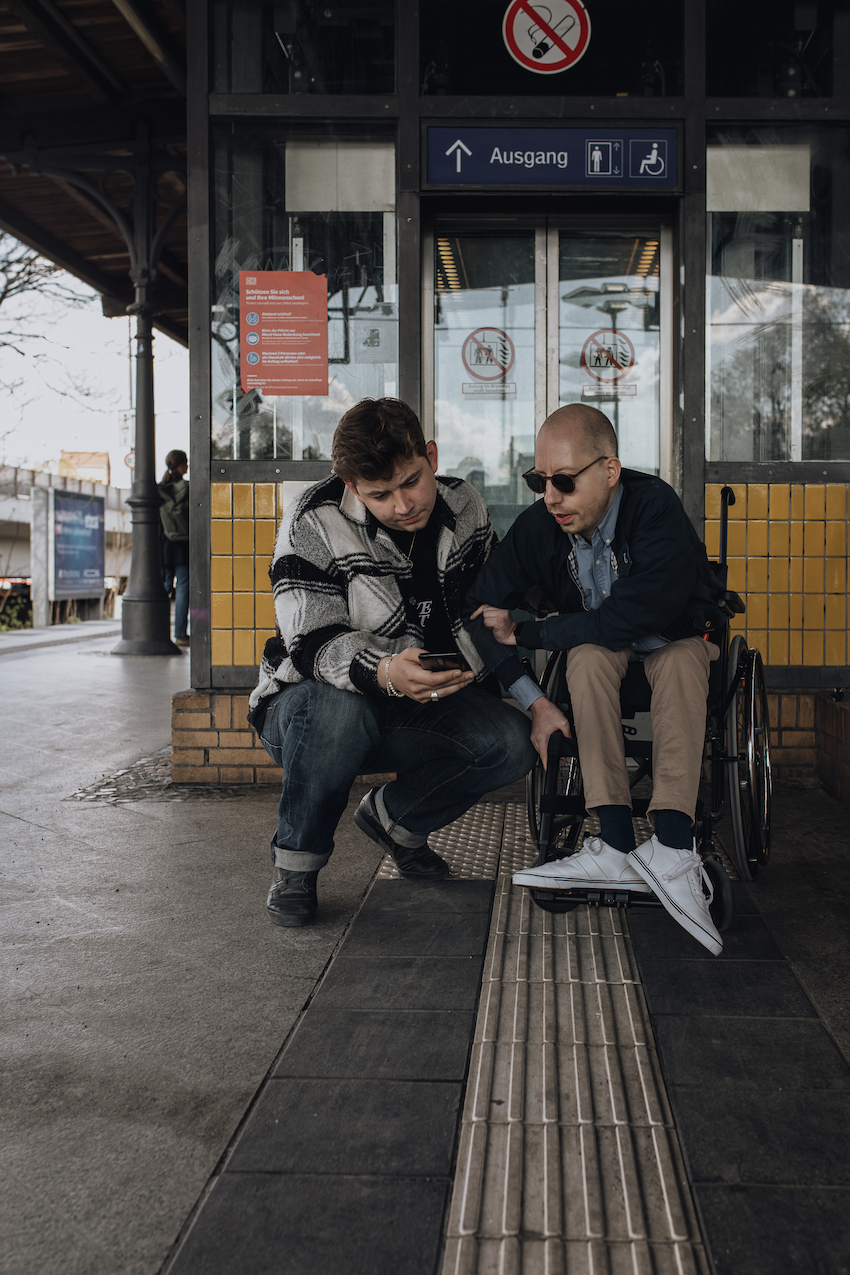 Foto: Auf einem Bahnsteig vor einem Aufzug sitzt ein Mann mit Sonnenbrille im Rollstuhl. Neben ihm kniet ein zweiter Mann. Beide betrachten ein Smartphone.