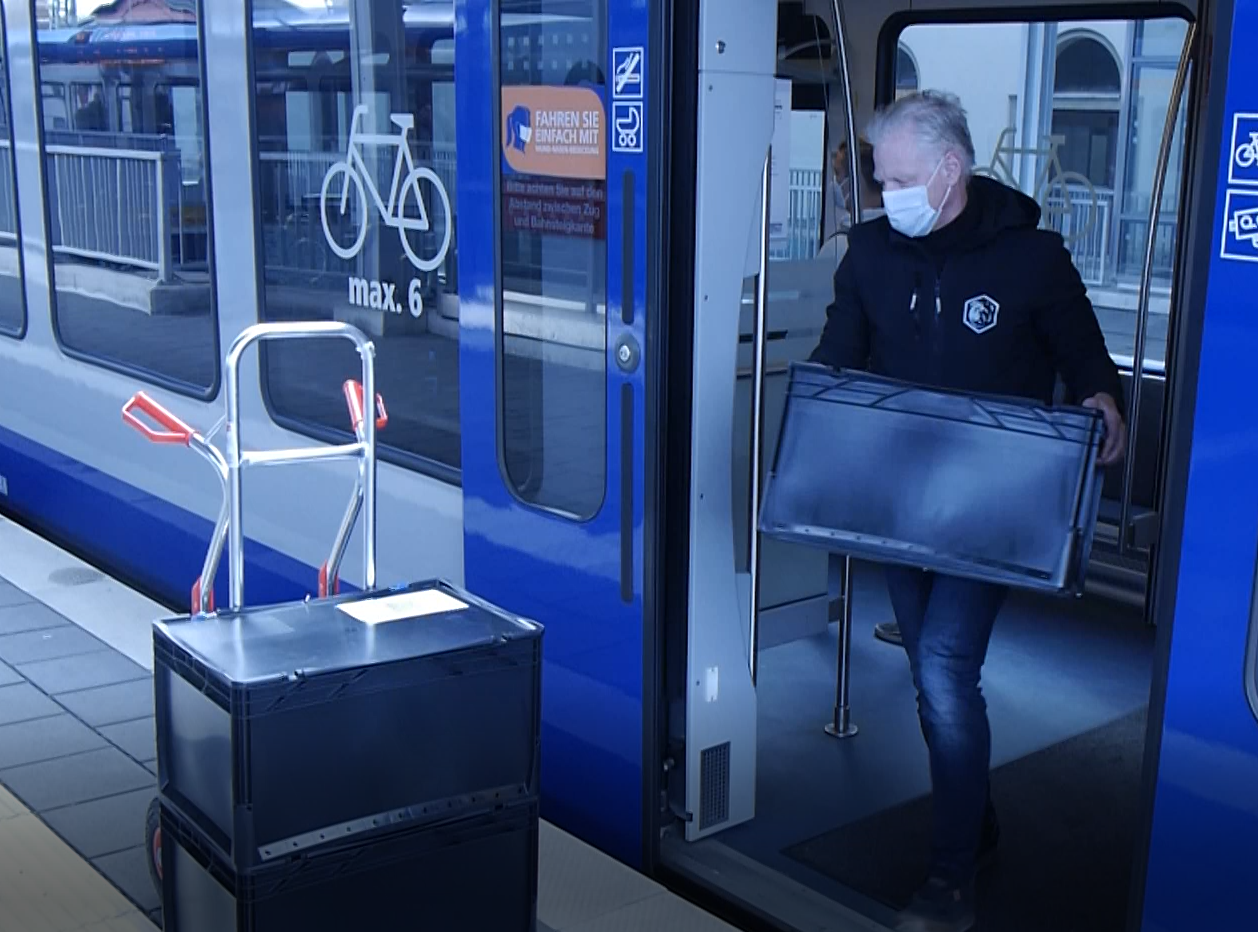 Foto: Ein Mann lädt eine große Kiste aus einer Straßenbahn auf eine Transportkarre auf dem Bahnsteig
