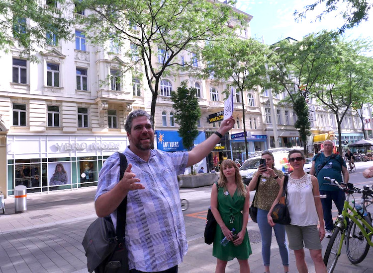 Mann im Karohemd steht in einer Fußgängerzone, erklärt etwas und hält ein Schild hoch, im Hintergrund stehen Menschen und hören ihm zu