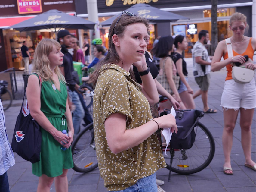 Seitliche Aufnahme einer Frau mit Zettel in der Hand in der Fußgängerzone, dahinter Umstehende und Passant*innen