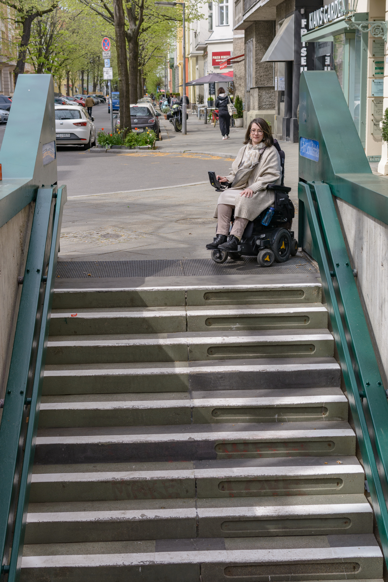 Eine Frau im Rollstuhl steht am oberen Rand einer langen Treppe, die abwärts z einem U-Bahnhof führt