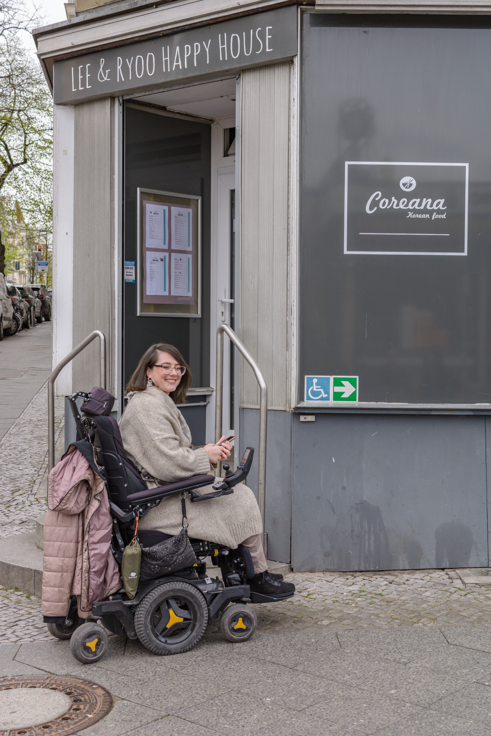 Eine Frau im Rollstuhl steht vor dem Eingang eines Restaurants, der eine hohe Schwelle hat. Große Piktogramme auf dem Fenster weisen ihr den Weg zum barrierefreien Eingang