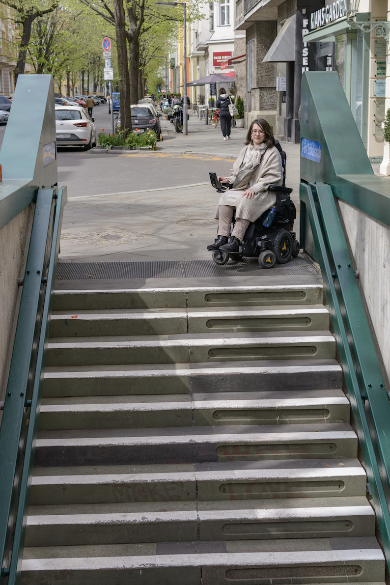 Eine Frau in einem elektrisch betriebenen Rollstuhl steht am oberen Ende einer Treppe, die hinab zu einem U-Bahnhof führt