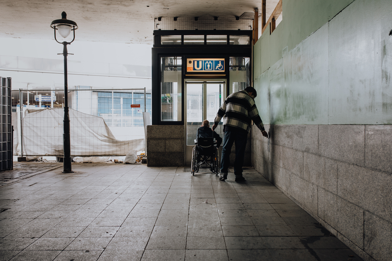 Ein Mann im Rollstuhl wartet mit seiner Assistenz vor einem Aufzug in einer düsteren Passage am U-Bahnhof