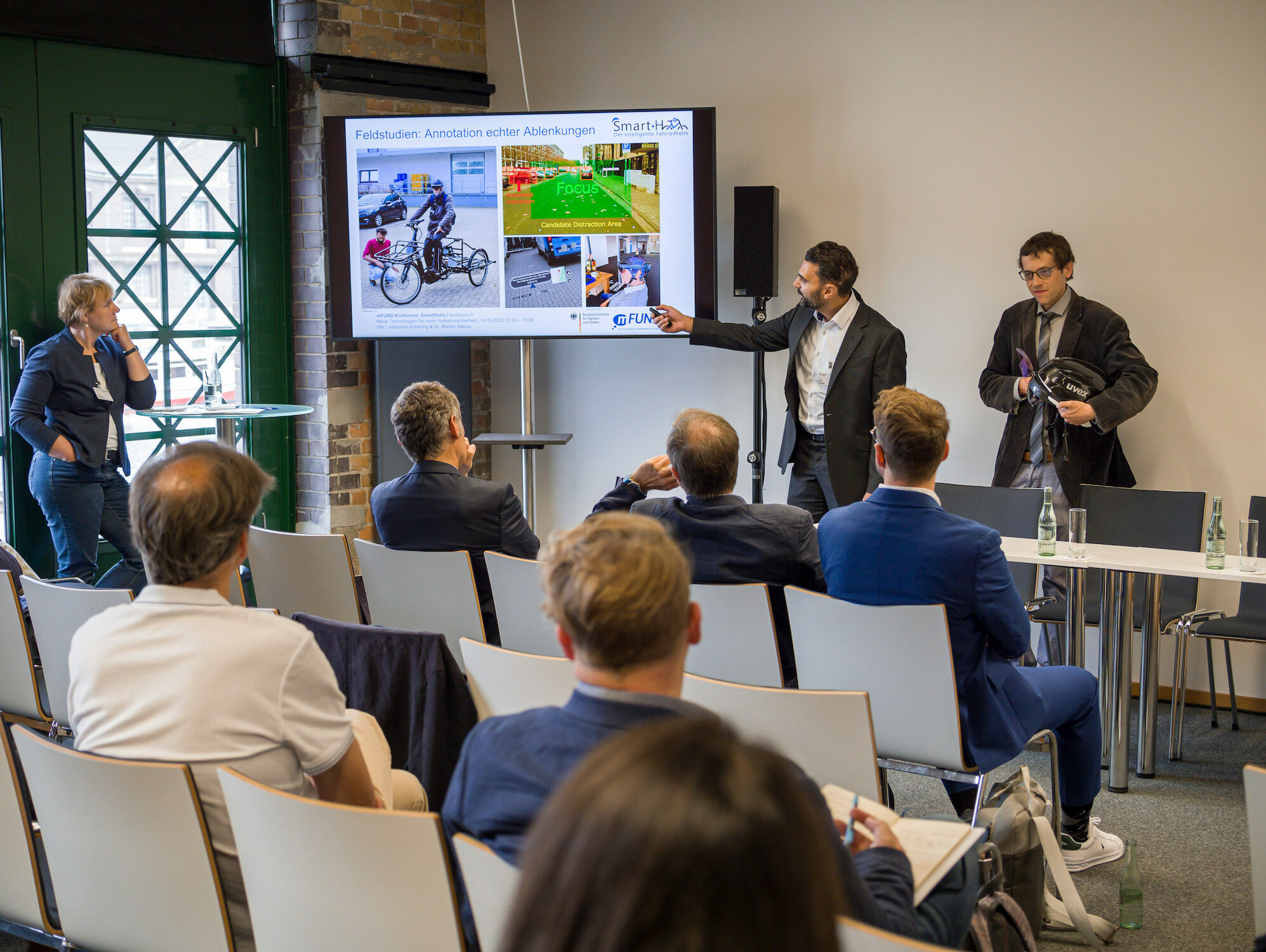 Neben einem großen Bildschirm stehen zwei Männer und erklären ihr Projekt SmartHelm dem Publikum im Raum